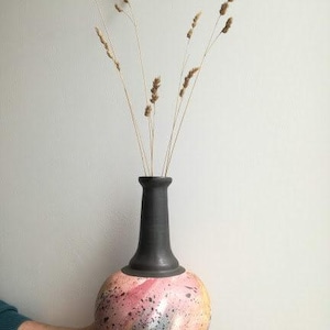 May include: A ceramic vase with a black neck and a round, speckled body. The body of the vase is painted with pink, yellow, and blue splatters. Dried wheat stalks are placed inside the vase.