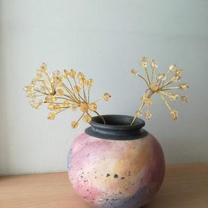 Könnte beinhalten: Eine Vase mit einem farbenfrohen, abstrakten Design hält zwei Zweige mit goldenen Perlenblumen. Die Vase ist in einem gesprenkelten, hellrosa, gelben und violetten Farbton gehalten.