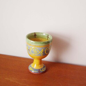 May include: A yellow ceramic goblet with a green rim and blue and yellow fish designs. The goblet has a short stem and a round base.