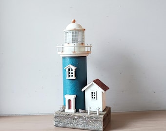 May include: A miniature lighthouse model with a teal blue tower, white accents, and a small attached white building with a red roof. The lighthouse sits on a wooden base, evoking a coastal theme.