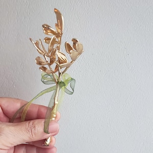 May include: A gold-coloured metal floral stem with multiple leaves and a green ribbon. The leaves have a smooth, reflective surface. The stem is held in a hand against a white background.