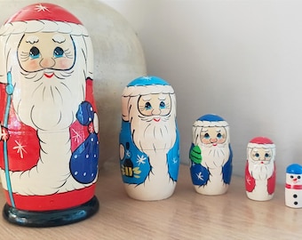 May include: Set of five traditional Russian nesting dolls, or matryoshka dolls, painted with Santa Claus designs. The largest doll is red and white with a blue bag, the next is blue and white, the next is blue and white with a green bag, the next is red and white, and the smallest is a snowman.