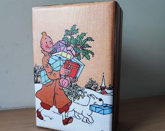 May include: A wooden block featuring a cartoon illustration of a person carrying gifts in a snowy scene. The person wears a brown coat and red shoes, accompanied by a white dog. The block has a light brown wood grain finish.