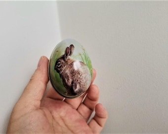 May include: A round, white, and green Easter egg-shaped tin with a brown rabbit illustration. The rabbit is facing left and has a white patch on its chest.
