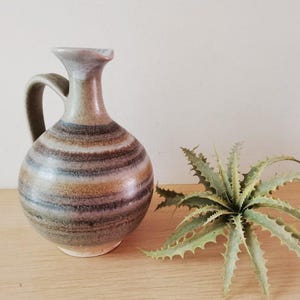 May include: A brown and grey striped ceramic jug with a wide mouth and a handle. The jug has a rustic, handmade look.