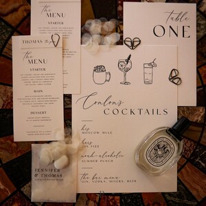 Può includere: Un insieme di articoli di cancelleria per matrimoni, tra cui menu, segnaposto e un menu bar, disposti su una superficie a motivi. I menu presentano un'elegante calligrafia e illustrazioni di cocktail. Sono presenti anche un flacone di profumo e fermagli decorativi.