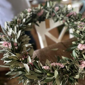 Puede incluir: Una corona floral hecha a mano con ramas de olivo y peque&ntilde;as flores rosas. La corona es circular y parece reci&eacute;n hecha, con hojas verdes y delicadas flores. Ideal para la decoraci&oacute;n del hogar o eventos especiales.