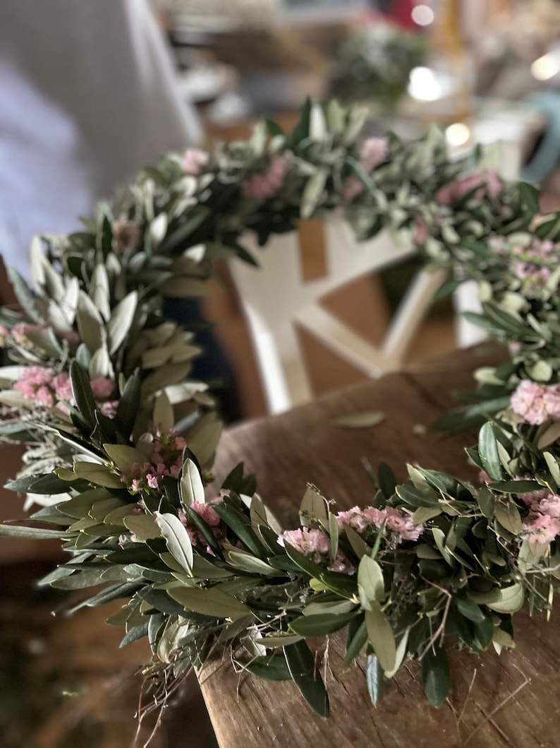 Puede incluir: Una corona floral hecha a mano con ramas de olivo y peque&ntilde;as flores rosas. La corona es circular y parece reci&eacute;n hecha, con hojas verdes y delicadas flores. Ideal para la decoraci&oacute;n del hogar o eventos especiales.