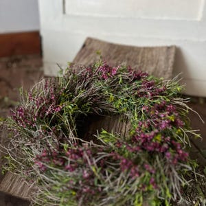 K&ouml;nnte beinhalten: Ein kreisf&ouml;rmiger Kranz aus d&uuml;nnen, getrockneten braunen Zweigen und &Auml;sten, der dicht mit kleinen, leuchtend magentafarbenen Bl&uuml;ten und zartem gr&uuml;nem Blattwerk durchwoben ist.