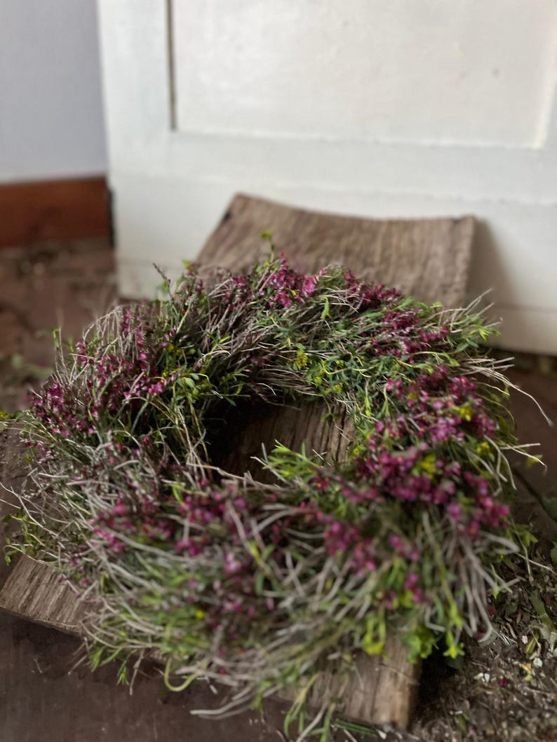 K&ouml;nnte beinhalten: Ein kreisf&ouml;rmiger Kranz aus d&uuml;nnen, getrockneten braunen Zweigen und &Auml;sten, der dicht mit kleinen, leuchtend magentafarbenen Bl&uuml;ten und zartem gr&uuml;nem Blattwerk durchwoben ist.