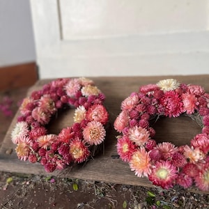 May include: Two circular wreaths made of dried flowers in shades of pink, red, and peach. The wreaths are arranged on a weathered wooden surface, with a white door in the background. The flowers are densely packed, creating a full, textured appearance.