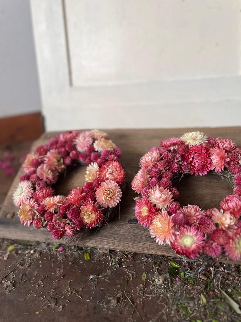 May include: Two circular wreaths made of dried flowers in shades of pink, red, and peach. The wreaths are arranged on a weathered wooden surface, with a white door in the background. The flowers are densely packed, creating a full, textured appearance.