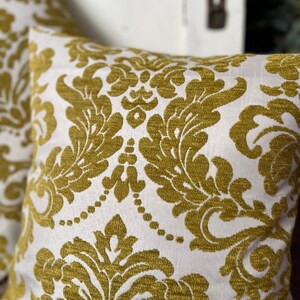 May include: Two square cushions featuring a white backdrop and a gold damask design. The cushions are made from a textured material, suitable for home decoration and adding a touch of elegance.