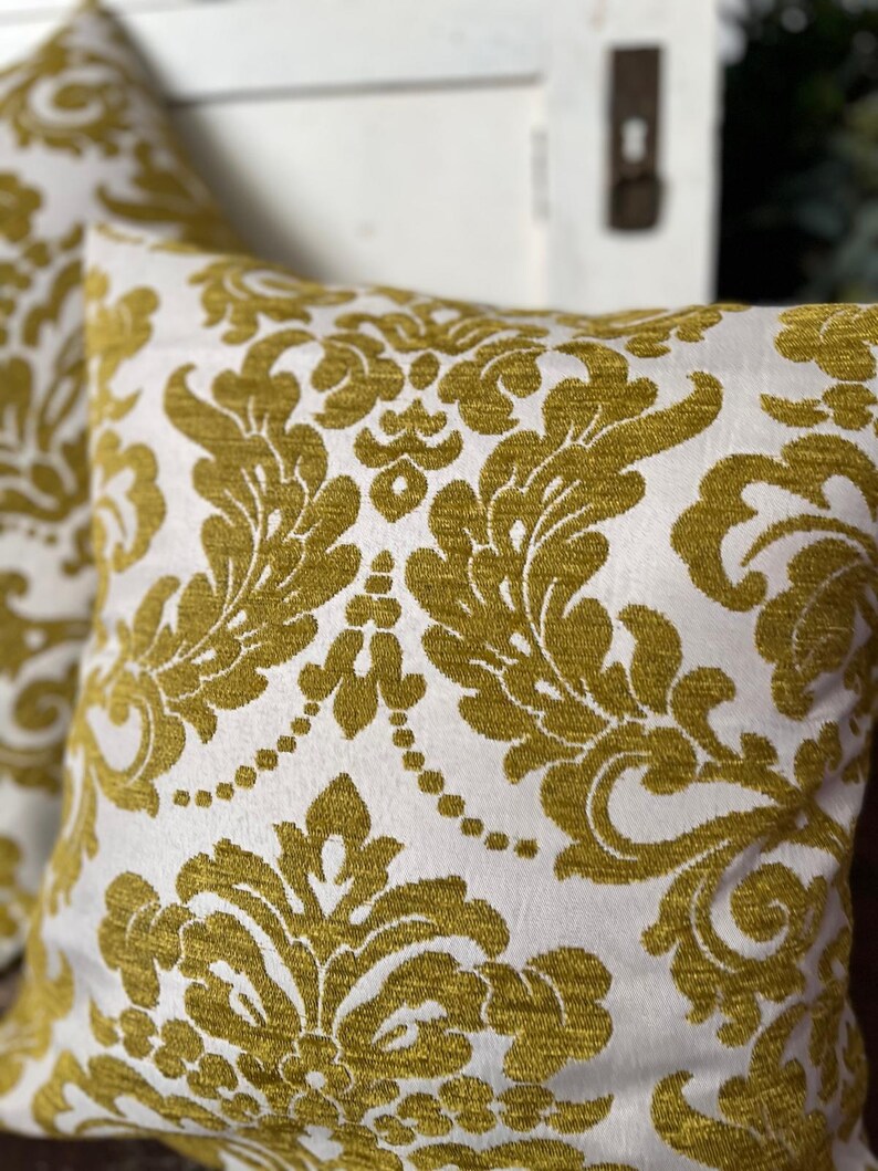 May include: Two square cushions featuring a white backdrop and a gold damask design. The cushions are made from a textured material, suitable for home decoration and adding a touch of elegance.