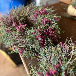 K&ouml;nnte beinhalten: Ein kreisf&ouml;rmiger Kranz aus d&uuml;nnen, holzigen braunen Zweigen und Stielen, dicht bedeckt mit kleinen, leuchtend magentafarbenen Bl&uuml;ten und gr&uuml;nem Laub. Das Arrangement hat ein strukturiertes, nat&uuml;rliches Aussehen mit vielen Stielen.
