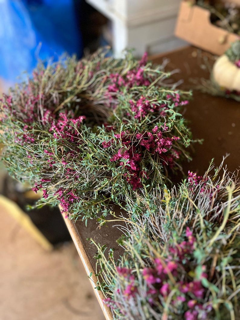 K&ouml;nnte beinhalten: Ein kreisf&ouml;rmiger Kranz aus d&uuml;nnen, holzigen braunen Zweigen und Stielen, dicht bedeckt mit kleinen, leuchtend magentafarbenen Bl&uuml;ten und gr&uuml;nem Laub. Das Arrangement hat ein strukturiertes, nat&uuml;rliches Aussehen mit vielen Stielen.