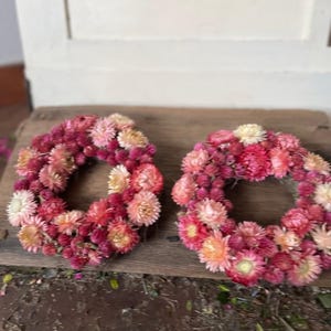May include: Two circular wreaths made of dried flowers in shades of pink, red, and cream. The wreaths are arranged on a weathered wooden surface, showcasing the natural textures and colors of the floral arrangement.