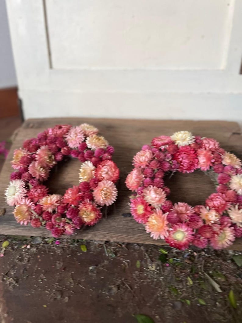 May include: Two circular wreaths made of dried flowers in shades of pink, red, and cream. The wreaths are arranged on a weathered wooden surface, showcasing the natural textures and colors of the floral arrangement.