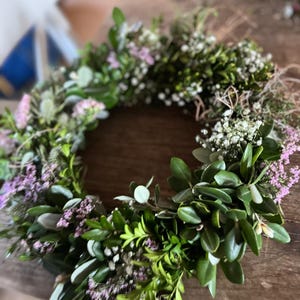 May include: A handmade floral wreath featuring a variety of greenery and small purple and white flowers. The wreath is circular and appears to be made with fresh or dried materials, perfect for home decor or special events.