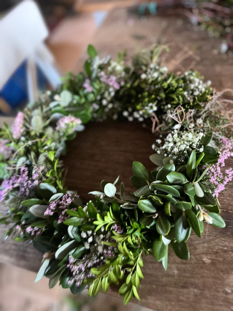 May include: A handmade floral wreath featuring a variety of greenery and small purple and white flowers. The wreath is circular and appears to be made with fresh or dried materials, perfect for home decor or special events.