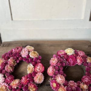 May include: Two floral wreaths composed of dried flowers in shades of pink, magenta, and cream. The wreaths are arranged on a weathered wooden surface, showcasing their circular form and natural textures.