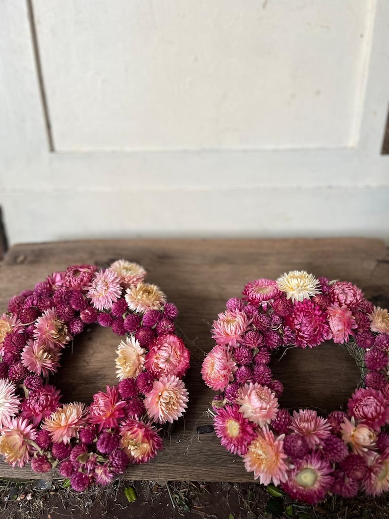 May include: Two floral wreaths composed of dried flowers in shades of pink, magenta, and cream. The wreaths are arranged on a weathered wooden surface, showcasing their circular form and natural textures.