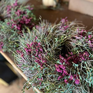 K&ouml;nnte beinhalten: Ein kreisf&ouml;rmiger Kranz aus getrockneten, d&uuml;nnen braunen Zweigen und St&auml;ngeln, dicht besetzt mit kleinen, leuchtend magentafarbenen Bl&uuml;ten und gr&uuml;nem Laub, angeordnet in einer nat&uuml;rlichen, strukturierten Komposition.