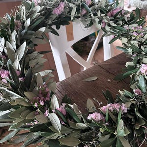 Puede incluir: Una corona hecha a mano con ramas de olivo y peque&ntilde;as flores rosas. La corona circular est&aacute; adornada con hojas y flores distribuidas uniformemente. Se encuentra sobre una mesa de madera.