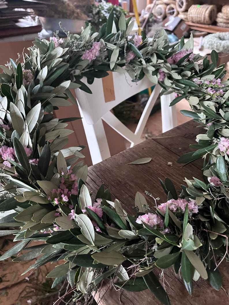 Puede incluir: Una corona hecha a mano con ramas de olivo y peque&ntilde;as flores rosas. La corona circular est&aacute; adornada con hojas y flores distribuidas uniformemente. Se encuentra sobre una mesa de madera.
