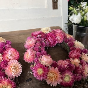 May include: Two floral wreaths made of dried flowers in shades of pink, peach, and cream. The wreaths are arranged on a weathered wooden surface. A door and a metal bucket with white flowers are in the background.