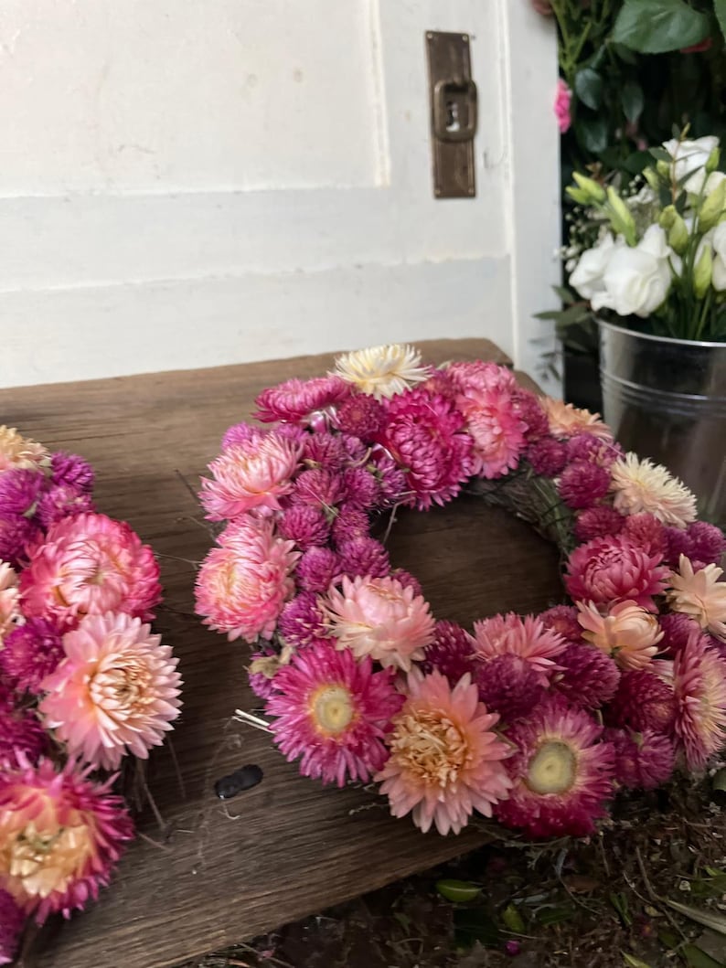 May include: Two floral wreaths made of dried flowers in shades of pink, peach, and cream. The wreaths are arranged on a weathered wooden surface. A door and a metal bucket with white flowers are in the background.