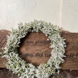 May include: A circular wreath of small white flowers and green leaves. The wreath is displayed on a wooden surface with engraved text. The design has a natural, rustic aesthetic.