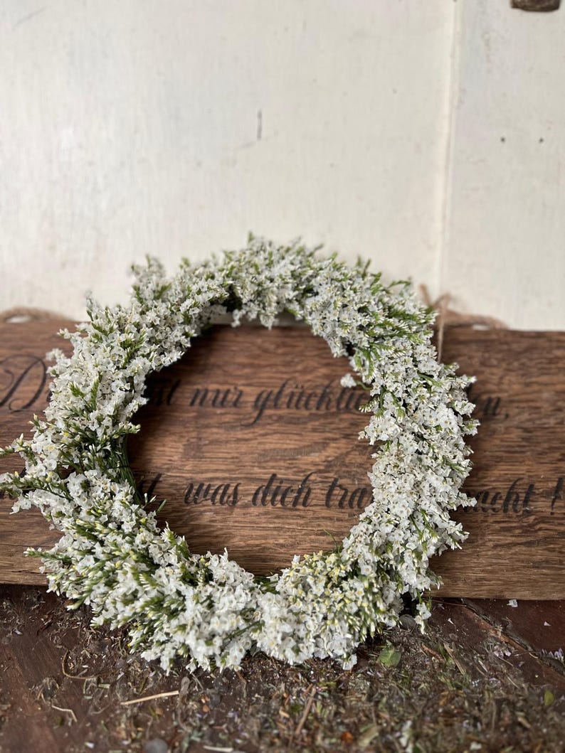 May include: A circular wreath of small white flowers and green leaves. The wreath is displayed on a wooden surface with engraved text. The design has a natural, rustic aesthetic.
