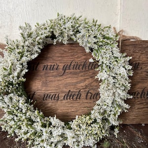 May include: A circular wreath of white flowers and green stems. The wreath is positioned in front of a wooden sign with engraved text. The sign is brown and has a natural wood grain. The text on the sign is in German.