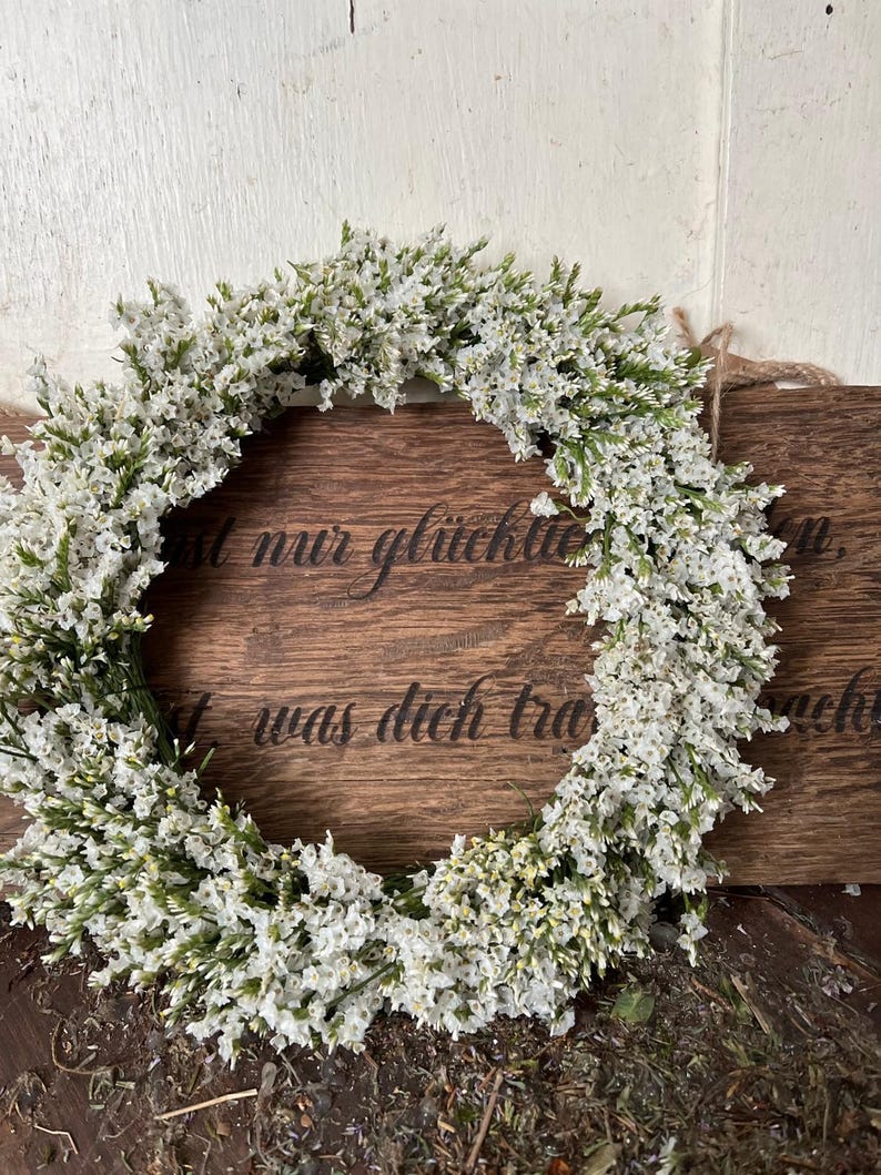 May include: A circular wreath of white flowers and green stems. The wreath is positioned in front of a wooden sign with engraved text. The sign is brown and has a natural wood grain. The text on the sign is in German.