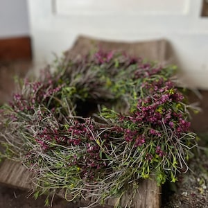 K&ouml;nnte beinhalten: Ein kreisf&ouml;rmiger Kranz aus d&uuml;nnen, getrockneten braunen Zweigen und &Auml;sten, dicht durchsetzt mit kleinen, leuchtend magentafarbenen Bl&uuml;ten und zartem gr&uuml;nem Laub, was ein strukturiertes, nat&uuml;rliches und abgerundetes botanisches Arrangement ergibt.