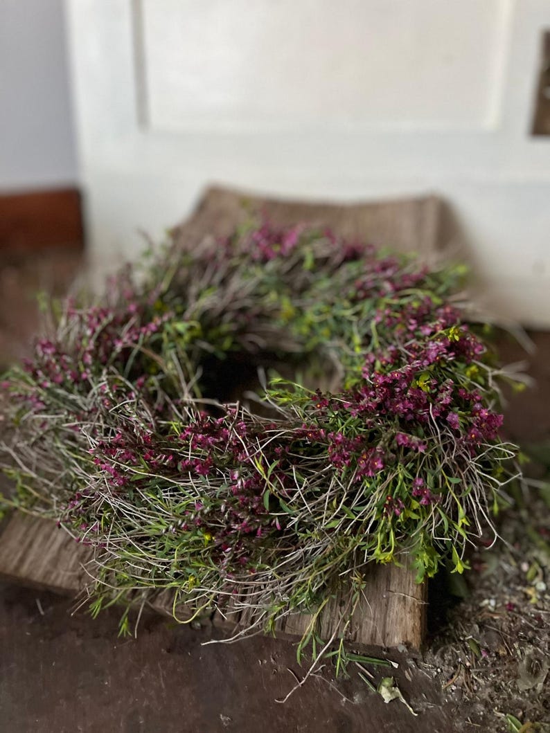K&ouml;nnte beinhalten: Ein kreisf&ouml;rmiger Kranz aus d&uuml;nnen, getrockneten braunen Zweigen und &Auml;sten, dicht durchsetzt mit kleinen, leuchtend magentafarbenen Bl&uuml;ten und zartem gr&uuml;nem Laub, was ein strukturiertes, nat&uuml;rliches und abgerundetes botanisches Arrangement ergibt.