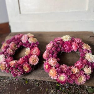 May include: Two circular wreaths made of dried flowers in shades of pink, magenta, and peach. The wreaths are arranged on a weathered wooden surface, showcasing the natural textures and colors of the floral arrangement.