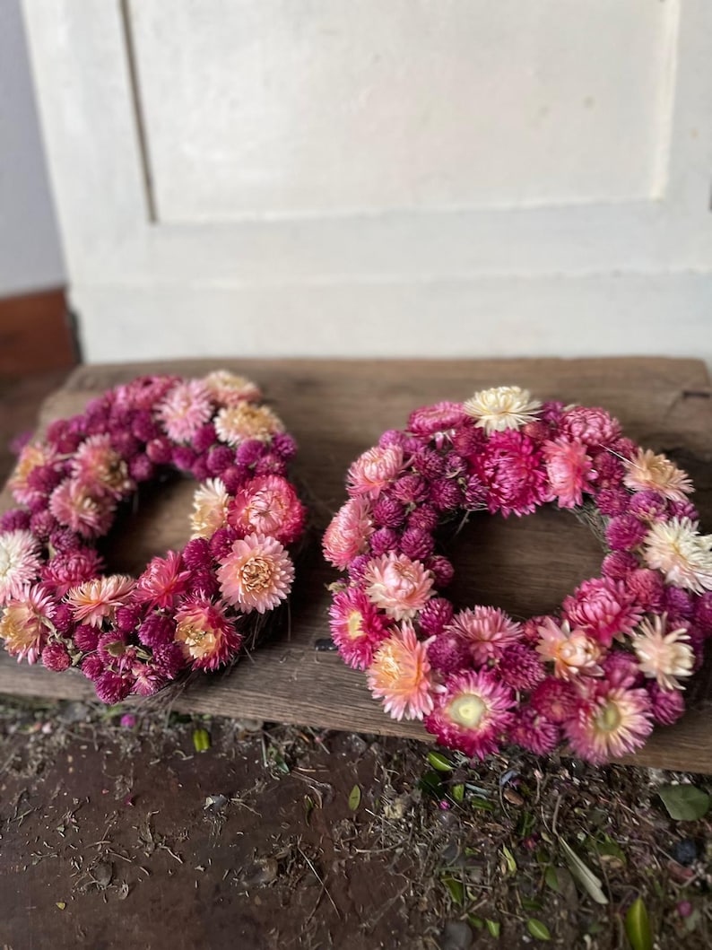 May include: Two circular wreaths made of dried flowers in shades of pink, magenta, and peach. The wreaths are arranged on a weathered wooden surface, showcasing the natural textures and colors of the floral arrangement.