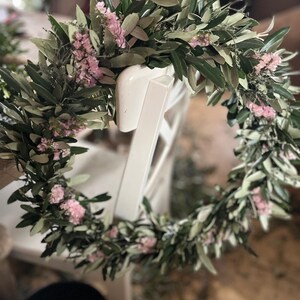 Puede incluir: Una corona floral circular compuesta por hojas verdes y peque&ntilde;as flores rosas, apoyada en el respaldo de una silla blanca. La corona est&aacute; hecha de vegetaci&oacute;n fresca y delicadas flores, creando un dise&ntilde;o natural y elegante.
