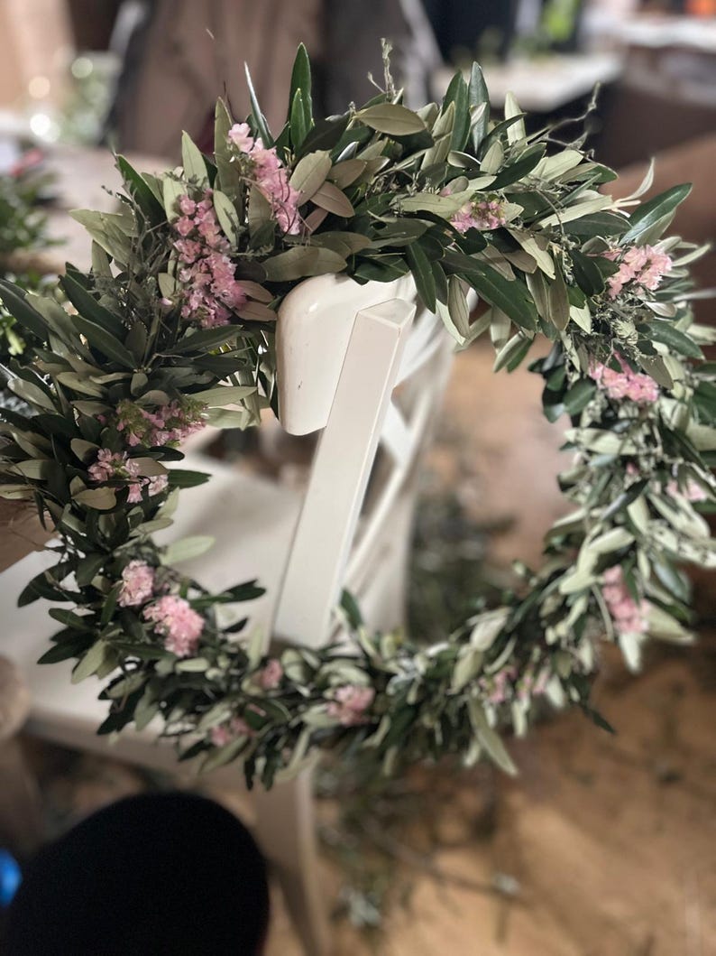 Puede incluir: Una corona floral circular compuesta por hojas verdes y peque&ntilde;as flores rosas, apoyada en el respaldo de una silla blanca. La corona est&aacute; hecha de vegetaci&oacute;n fresca y delicadas flores, creando un dise&ntilde;o natural y elegante.