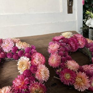 May include: Two floral wreaths composed of pink, magenta, and peach-colored dried flowers. The wreaths are arranged on a wooden surface, with a white door and a glimpse of other flowers in the background. The flowers are densely packed, creating a full, textured appearance.