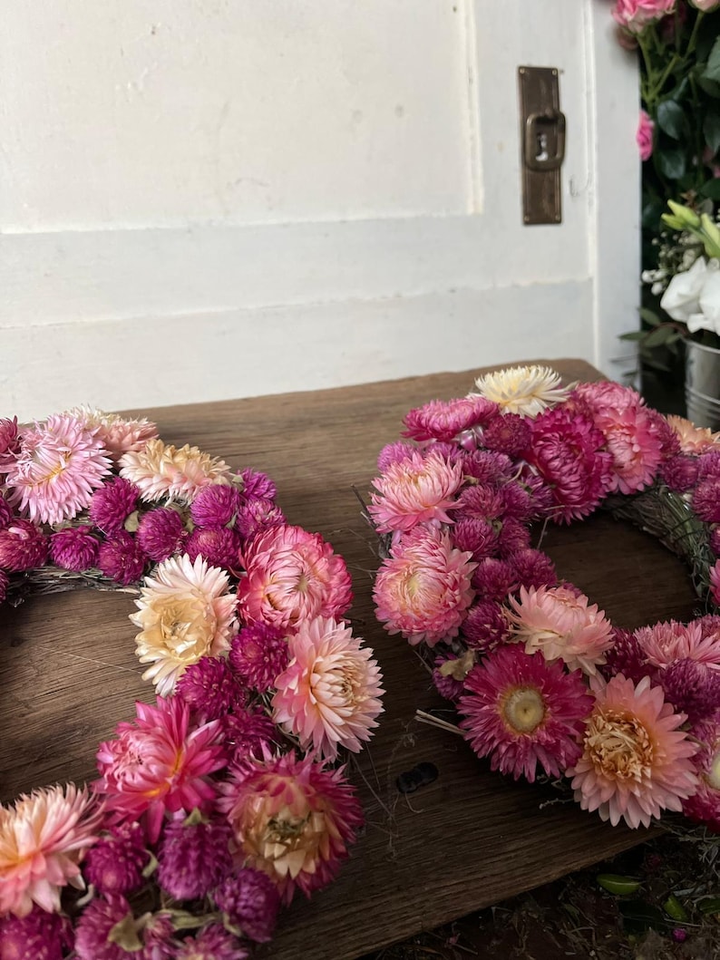 May include: Two floral wreaths composed of pink, magenta, and peach-colored dried flowers. The wreaths are arranged on a wooden surface, with a white door and a glimpse of other flowers in the background. The flowers are densely packed, creating a full, textured appearance.