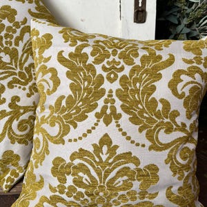 May include: Two square cushions featuring a white backdrop and a gold damask design. The cushions are made from a textured material and are positioned on a wooden surface. These cushions are a decorative home decor item.