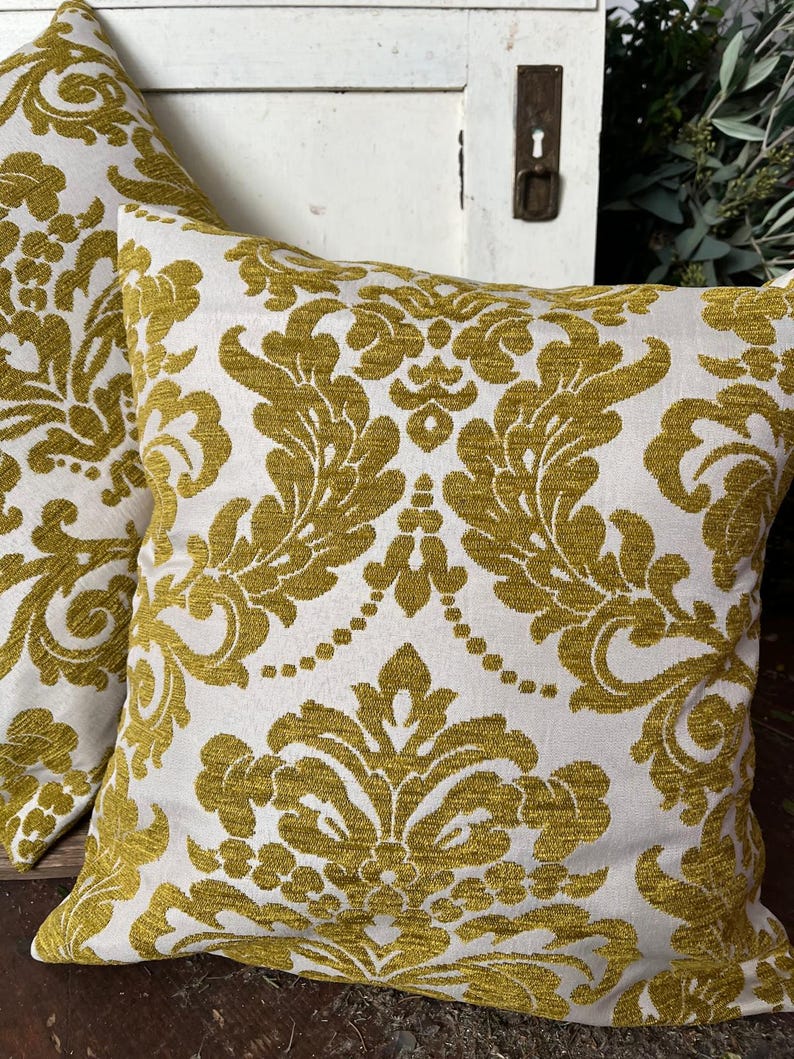 May include: Two square cushions featuring a white backdrop and a gold damask design. The cushions are made from a textured material and are positioned on a wooden surface. These cushions are a decorative home decor item.