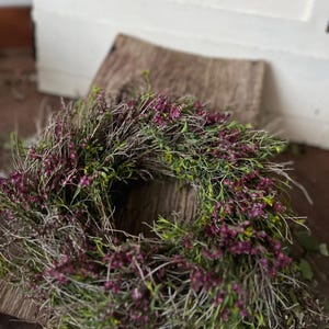 K&ouml;nnte beinhalten: Ein kreisf&ouml;rmiger Kranz aus d&uuml;nnen, getrockneten braunen Zweigen und &Auml;sten, dicht verwoben mit kleinen, leuchtend violetten, heidekrautartigen Bl&uuml;ten und feinem gr&uuml;nen Blattwerk, was ein strukturiertes, nat&uuml;rliches Aussehen ergibt.