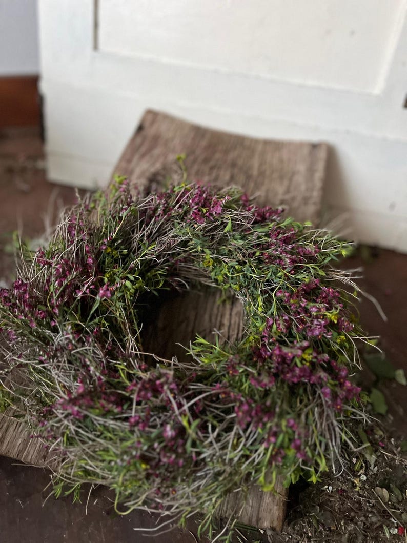 K&ouml;nnte beinhalten: Ein kreisf&ouml;rmiger Kranz aus d&uuml;nnen, getrockneten braunen Zweigen und &Auml;sten, dicht verwoben mit kleinen, leuchtend violetten, heidekrautartigen Bl&uuml;ten und feinem gr&uuml;nen Blattwerk, was ein strukturiertes, nat&uuml;rliches Aussehen ergibt.