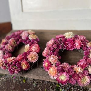 May include: Two floral wreaths composed of pink, magenta, and peach-colored dried flowers. The wreaths are circular and rest on a wooden surface. The flowers are densely packed, creating a full, textured appearance. The wreaths are a decorative item.