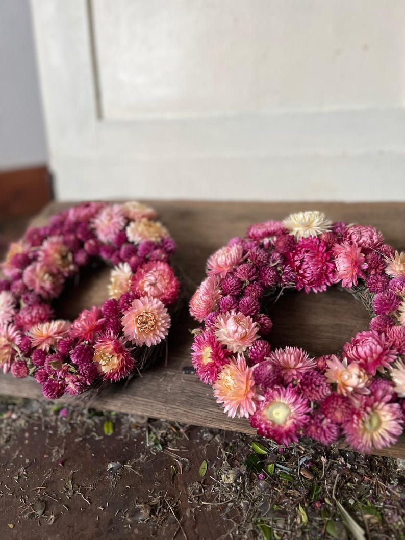 May include: Two floral wreaths composed of pink, magenta, and peach-colored dried flowers. The wreaths are circular and rest on a wooden surface. The flowers are densely packed, creating a full, textured appearance. The wreaths are a decorative item.