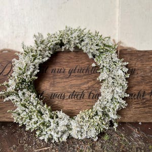 May include: A circular wreath composed of small white flowers and green stems. The wreath is presented on a wooden surface with a wooden sign in the background. The sign has text on it.